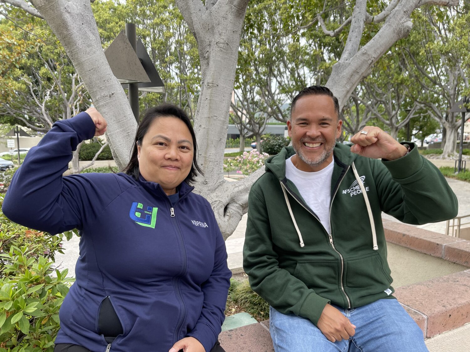 two nurses sit outside behind trees with fists up and smile