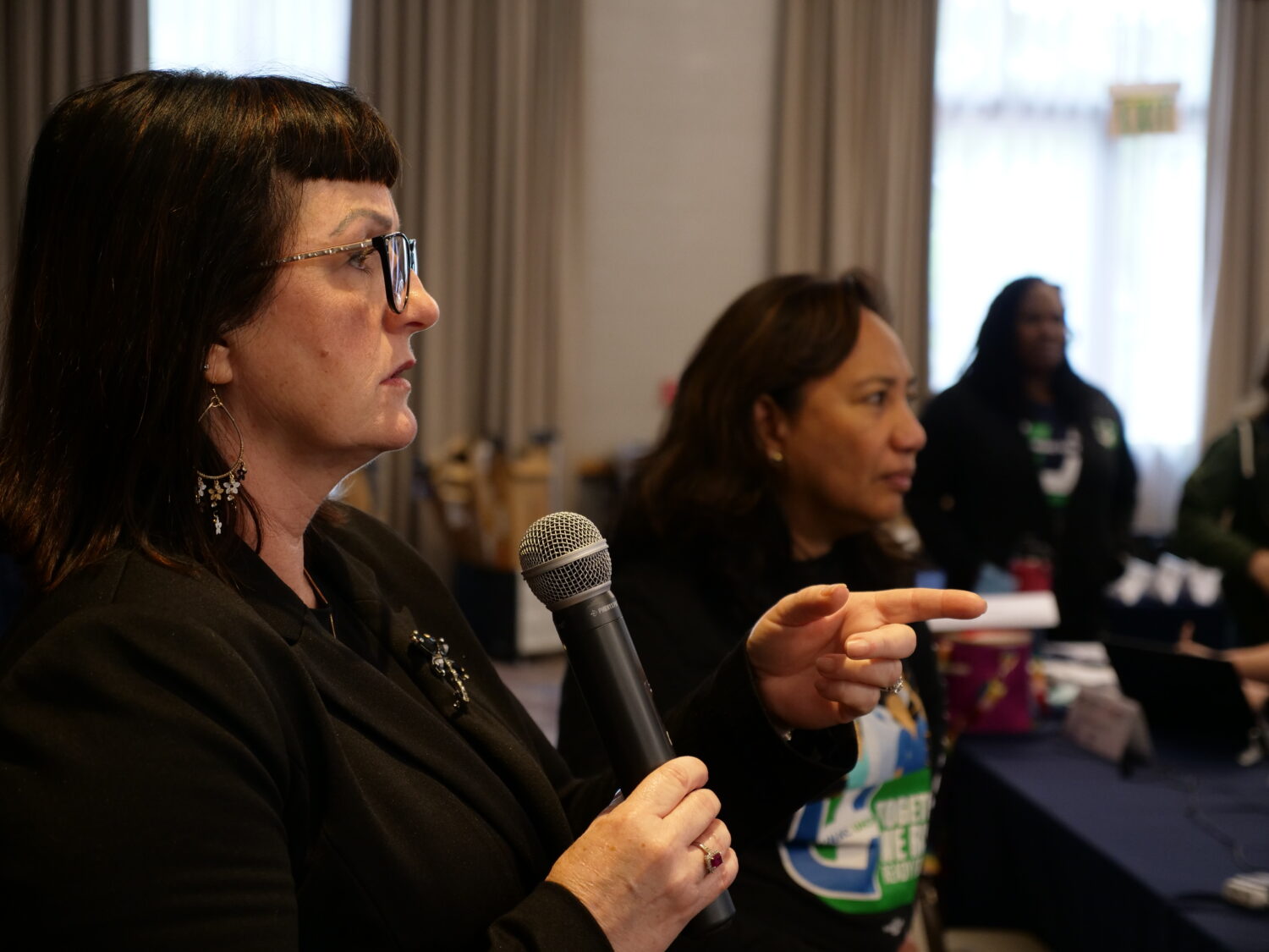woman speaks into mic seriously with others in background listening